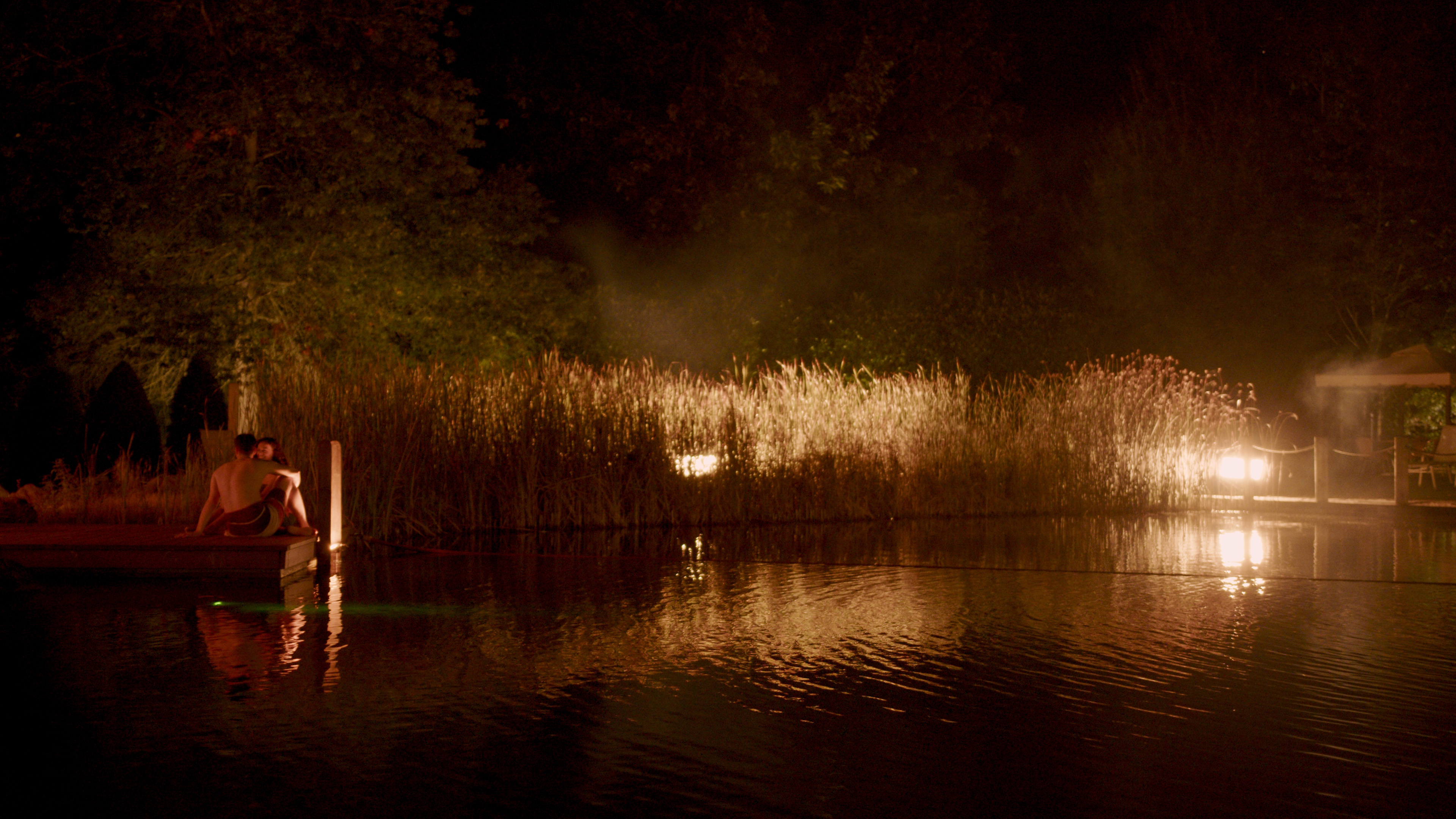 Das Bild zeigt eine friedliche Nachtszene am Wasser, umgeben von Bäumen und sanft beleuchtet von einigen Lichtern. 