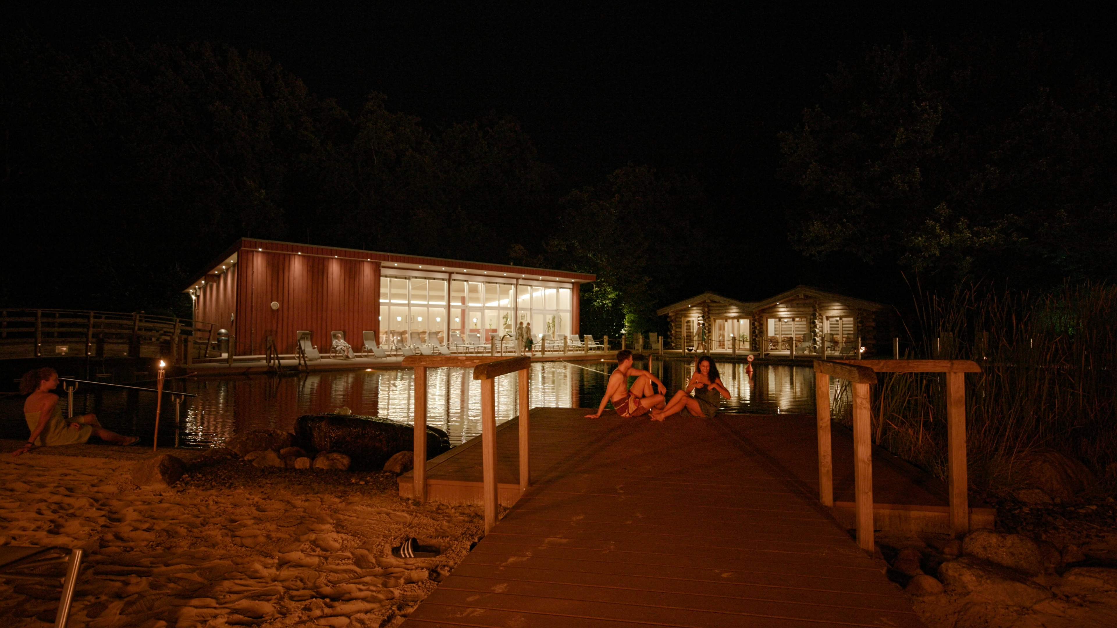 Nachtansicht einer beleuchteten Wellness-Anlage. Das Bild zeigt eine beleuchtete Wellness- oder Spa-Anlage in der Nacht, die von einem Holzsteg aus sichtbar ist.