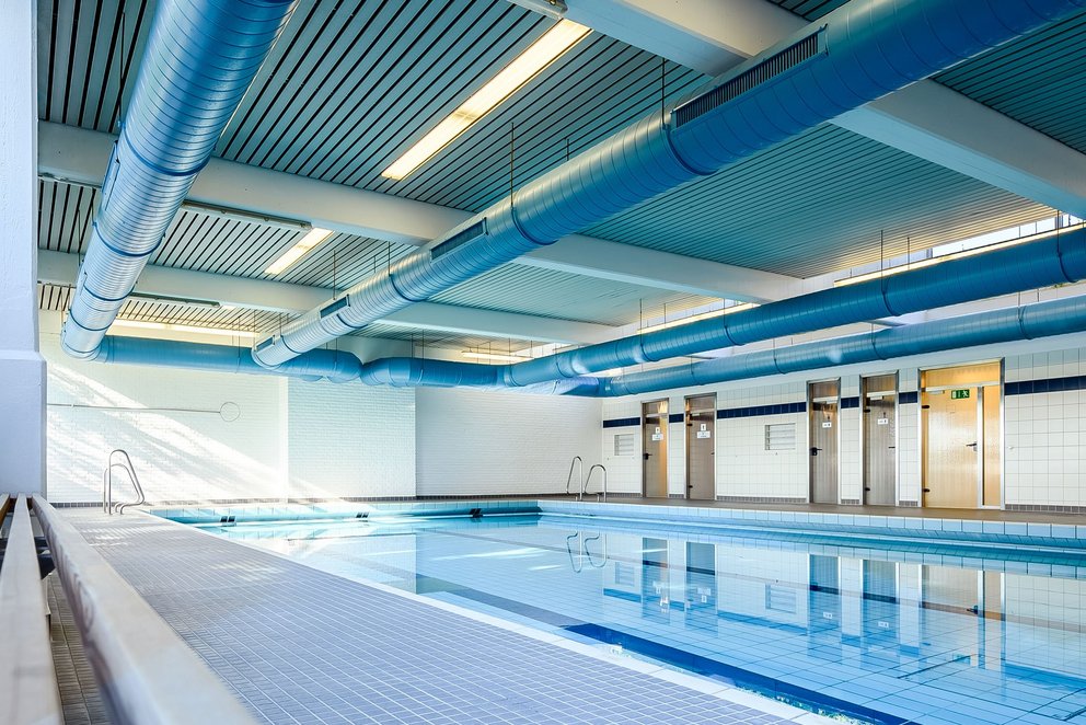 Modernes Hallenbad mit Holzbank. Das Bild zeigt einen modernen Hallenbadbereich mit einem klaren Schwimmbecken und einer eleganten Holzbank am Rand. Die Architektur mit geschwungener Decke und viel natürlichem Licht schafft eine angenehme und offene Atmosphäre.
