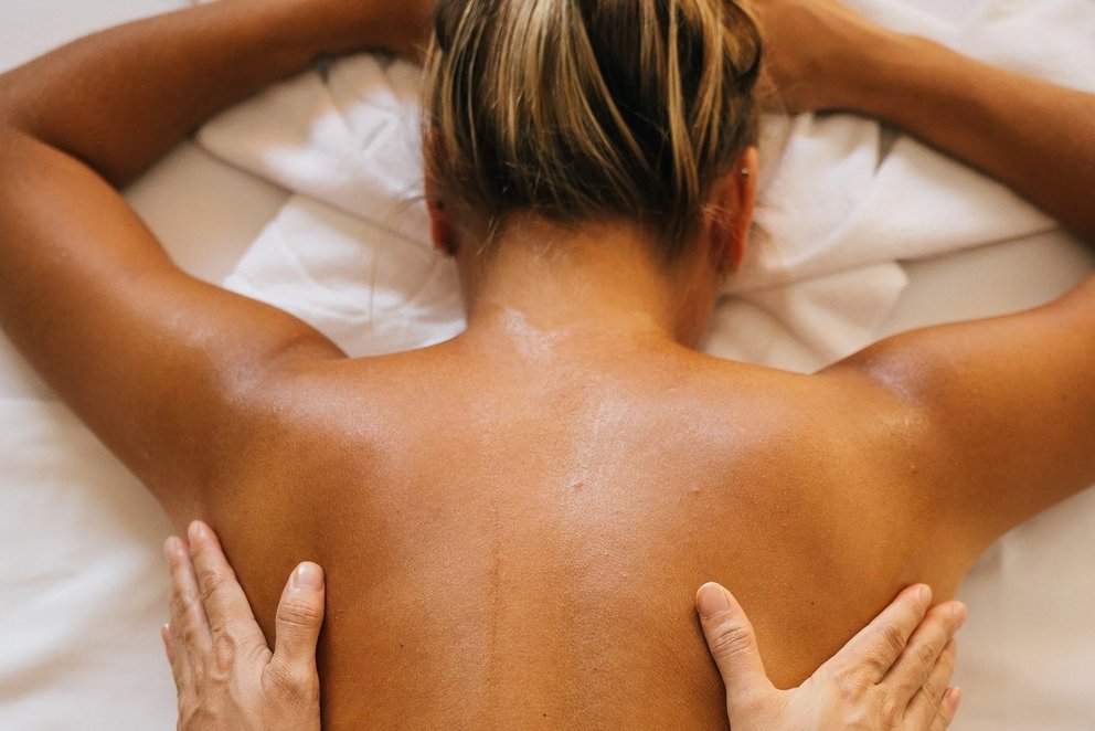Person erhält entspannende Massage am Nacken. Das Bild zeigt eine Person, die eine entspannende Massage erhält. Sie liegt auf dem Bauch, mit einem Handtuch umwickelt, und ein Masseur arbeitet sanft an ihrem Nacken- und Schulterbereich.