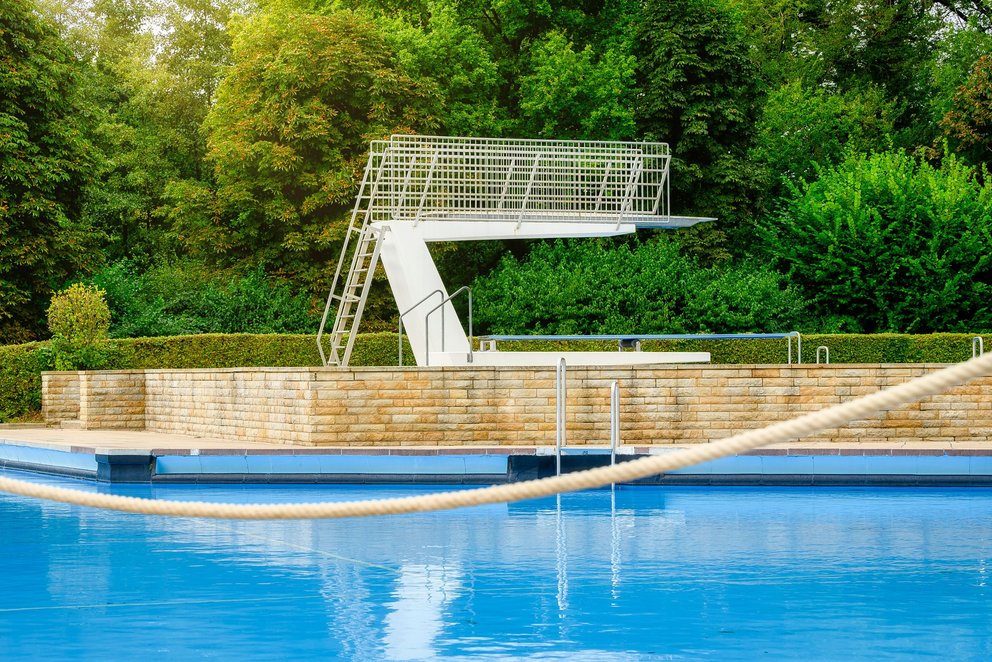 Das Bild zeigt ein Sprungbecken mit einem Sprungturm, umgeben von grünen Bäumen. Das klare Wasser und die sonnige Atmosphäre machen es zu einem idealen Ort für Sprungtraining und Erholung.