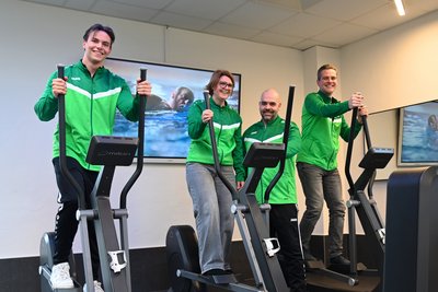 Das Bild zeigt vier Personen, die auf Ellipsentrainern in einem Fitnessstudio trainieren. Alle tragen grüne Sportjacken und strahlen eine positive, motivierte Energie aus.