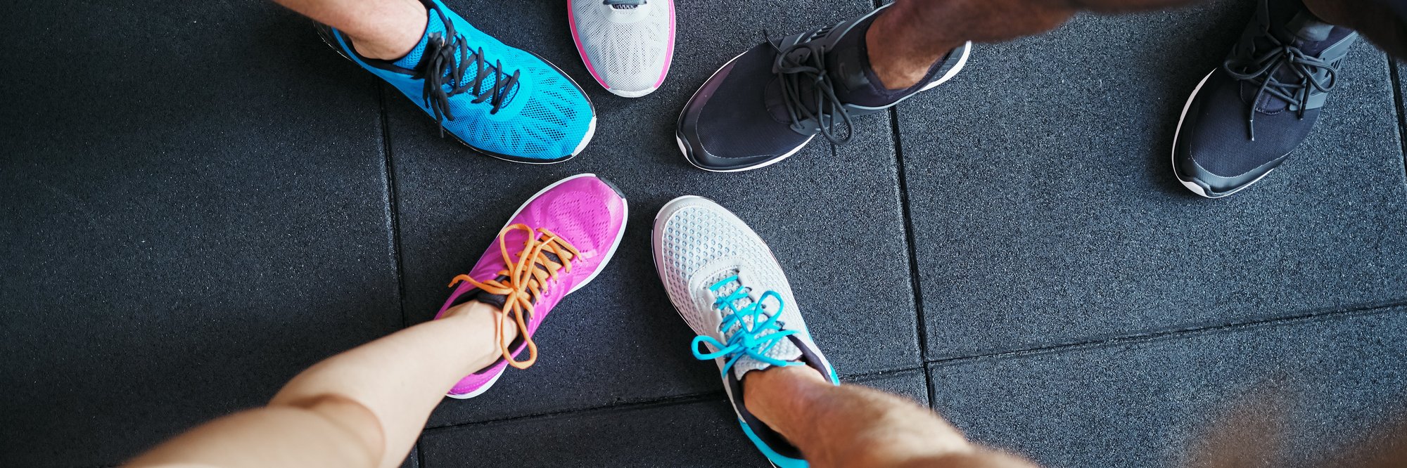 Vier Paare bunter Sportschuhe im Kreis. Das Bild zeigt vier Paare von Füßen, die im Kreis angeordnet sind. Jeder trägt bunte Sportschuhe, und der Boden besteht aus schwarzem, strukturiertem Material.