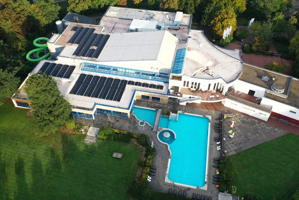 Luftaufnahme eines Schwimmbads. Das Bild zeigt eine Luftaufnahme eines Schwimmbads mit Innen- und Außenbereich. Zu sehen sind ein großes Freibadbecken, Rutschen, Sonnenterrassen und umgebendes Grün – eine vielfältige Anlage für Sport und Erholung.