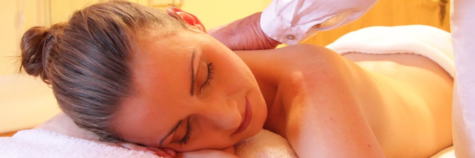 Person erhält entspannende Massage am Nacken. Das Bild zeigt eine Person, die eine entspannende Massage erhält. Sie liegt auf dem Bauch, mit einem Handtuch umwickelt, und ein Masseur arbeitet sanft an ihrem Nacken- und Schulterbereich.