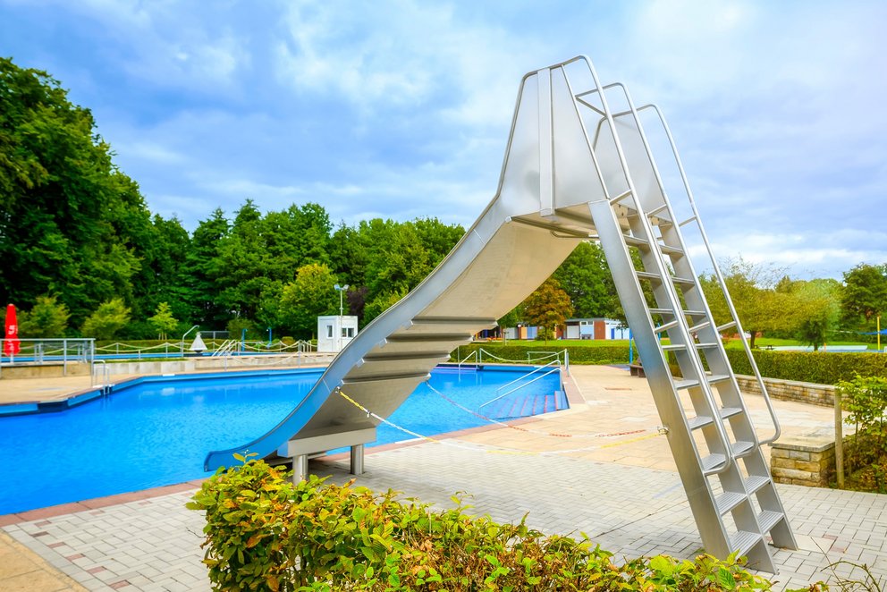 Das Bild zeigt eine Rutsche neben einem Freibadbecken, umgeben von grünen Büschen und einem weiten Rasenbereich. Die sonnige Atmosphäre macht den Ort einladend für Spaß und Erholung im Freien.