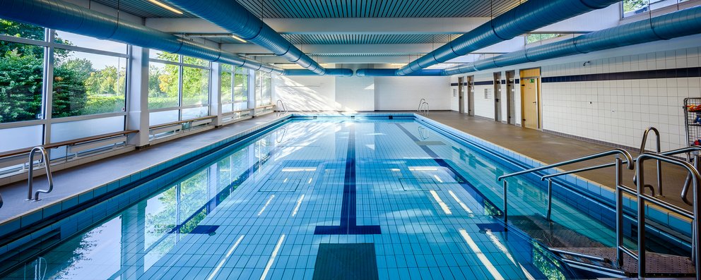 Hallenbad mit langen Schwimmbahnen. Das Bild zeigt ein Hallenbad.Durch die großen Fenster dringt viel Tageslicht in den Raum, was eine helle und einladende Atmosphäre schafft.
