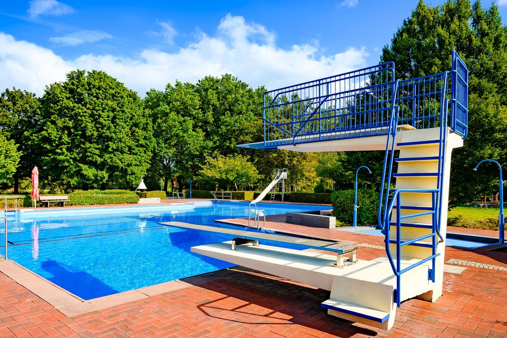 Das Bild zeigt ein Freibad mit Sprungturm vor grünem Baumbestand. Das klare Wasser, die rote Pflasterung und der blaue Himmel schaffen eine einladende Atmosphäre für Sommer, Spaß und sportliche Aktivitäten.