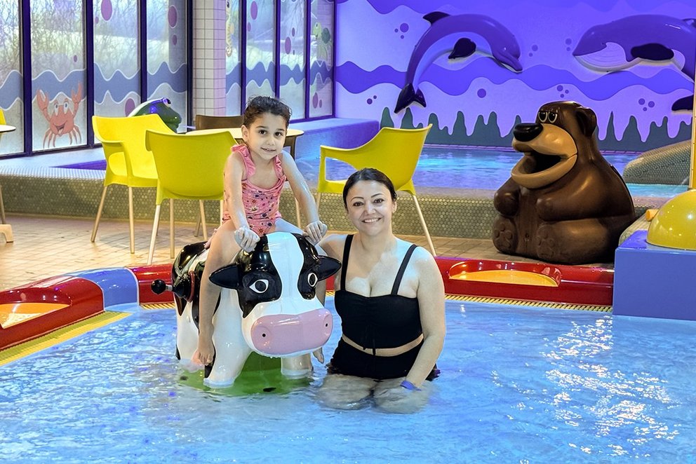 Das Bild zeigt eine Mutter und ihre Tochter, die zusammen in einem Kinderbecken in einem Schwimmbad stehen. 