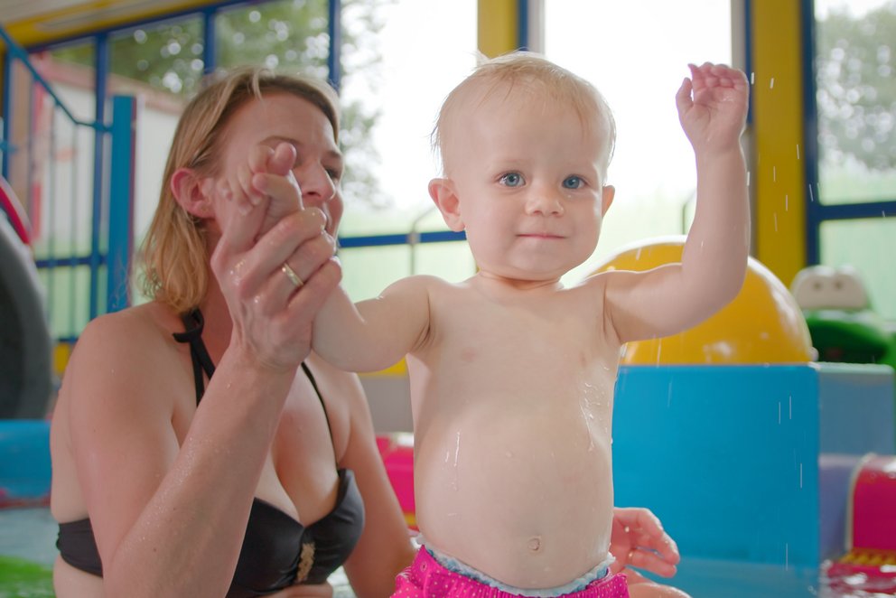 Mutter unterstützt Kind im Hallenbad. Das Bild zeigt eine Mutter, die ihr kleines Kind im Wasser eines Hallenbads unterstützt. Das Kind steht im Wasser und hebt freudig die Arme, während die Mutter es sicher festhält.
