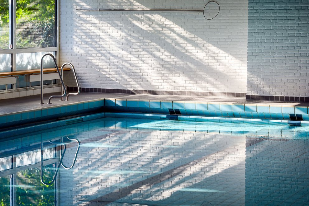 Innenpool mit sanftem Licht und klarer Wasseroberfläche. Das Bild zeigt einen ruhigen Teil eines Hallenbades. Das klare Wasser spiegelt das Licht, das durch die Fenster auf die Pooloberfläche fällt. Die Atmosphäre ist ruhig und einladend, ideal zum Entspannen.