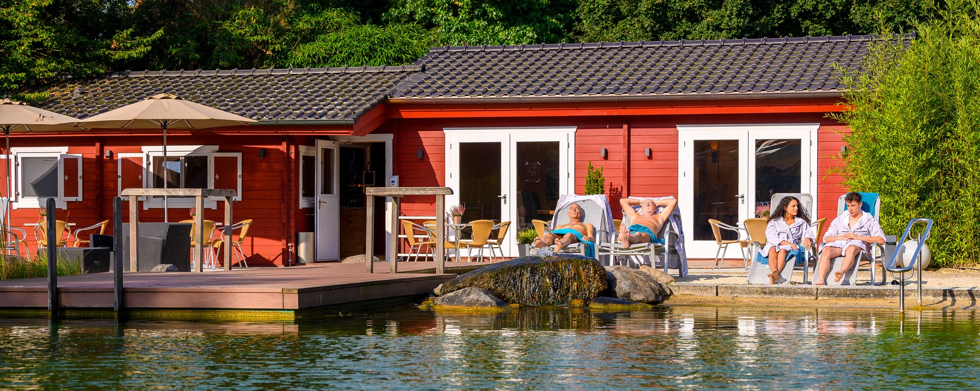 Haus am Wasser mit Steg und grüner Umgebung. Rotes Haus am Wasser mit einem Steg, umgeben von ruhigem Wasser und grüner Vegetation im Hintergrund.