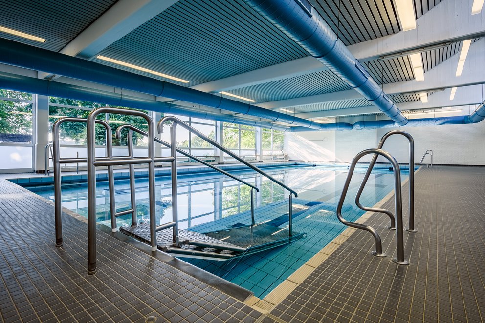 Zugang zum Schwimmbecken im modernen Hallenbad. Das Bild zeigt den Zugang zu einem Schwimmbecken in einem modernen Hallenbad. Mit eleganten Metallgeländern und einer großzügigen, hellen Architektur wirkt der Raum offen und einladend. Viel Tageslicht durchflutet den Raum und sorgt für eine angenehme Atmosphäre.