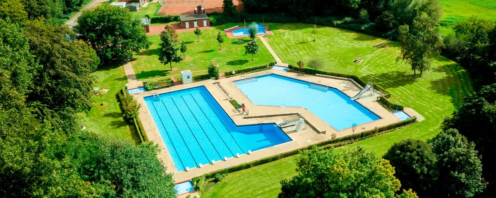 Das Bild zeigt ein malerisches Freibad aus der Hochperspektive, mit zwei Becken, die durch eine kleine Brücke miteinander verbunden sind. Umgeben von viel Grün und Bäumen bietet die Anlage eine ruhige und entspannte Atmosphäre.