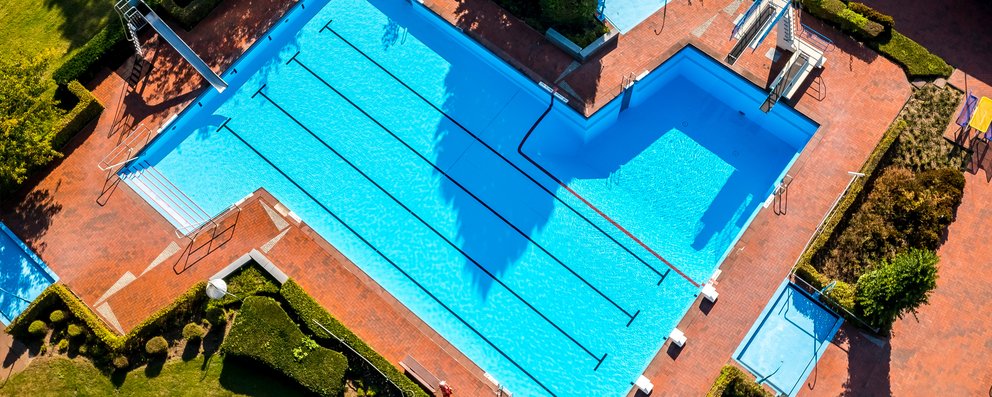Das Bild zeigt ein großes Freibad aus der Vogelperspektive. Die Becken sind klar strukturiert mit Schwimmbahnen, Sprungbereich und einem flachen Bereich – umgeben von Bäumen und roten Pflasterwegen. Eine gepflegte Anlage für Sport, Spaß und Erholung.